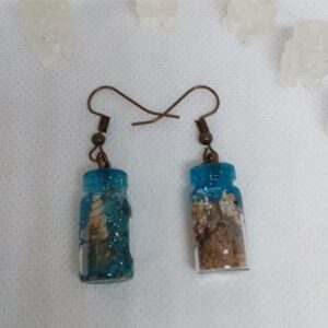 Handcrafted resin bottle earrings filled with real sand and shells, photographed on a coastal shore in warm cinematic light.